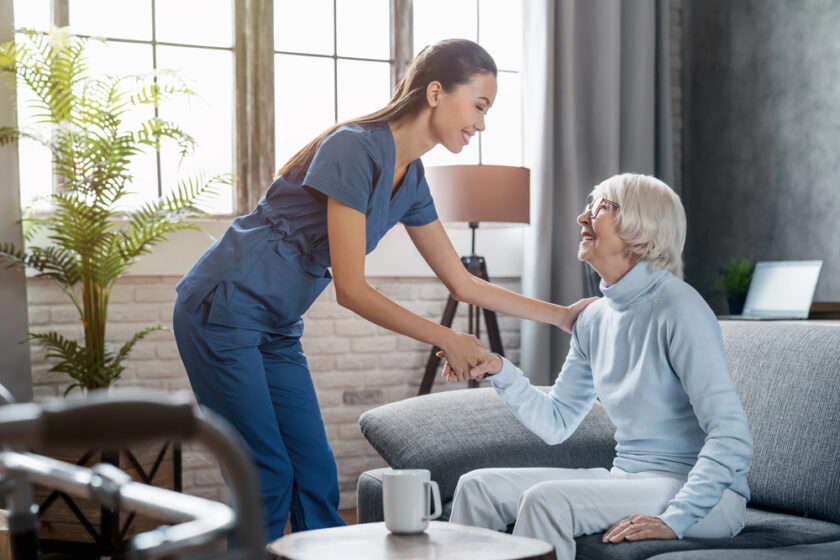 A nurse in blue scrubs smiles and holds hands with a seated elderly woman, who is also smiling, in a bright, cosy living room with large windows and plants.