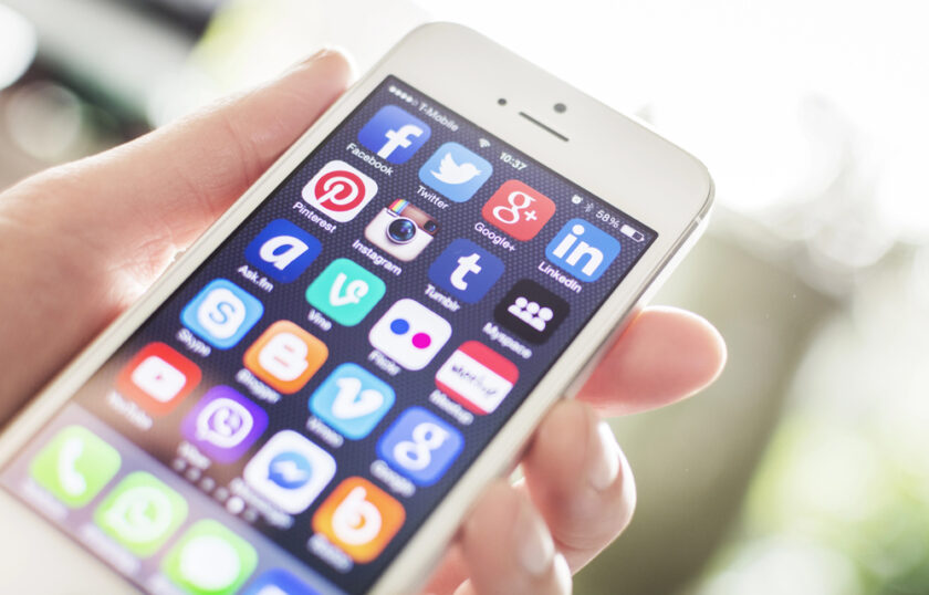 A hand holding a white mobile phone displaying various social media app icons on the screen, including Facebook, Twitter, Google+, LinkedIn, Pinterest, Instagram, and others.