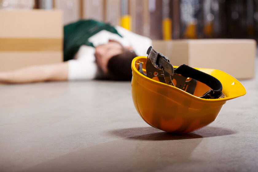 A yellow safety helmet lies on the floor in focus, with a person wearing work clothes lying motionless and blurred in the background, suggesting a workplace accident or injury.