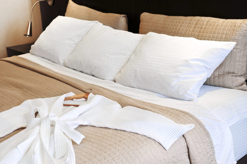 A neatly made bed with white pillows and beige bedding. A white bathrobe is laid out on top of the bed with a wooden hanger, next to a reading lamp.