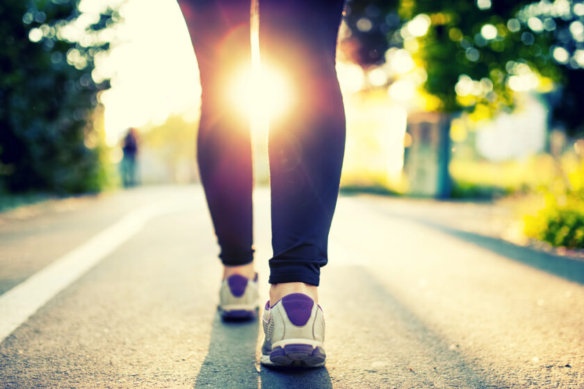A person wearing trainers and black leggings walks on a path outdoors with sunlight shining brightly through the trees in the background.
