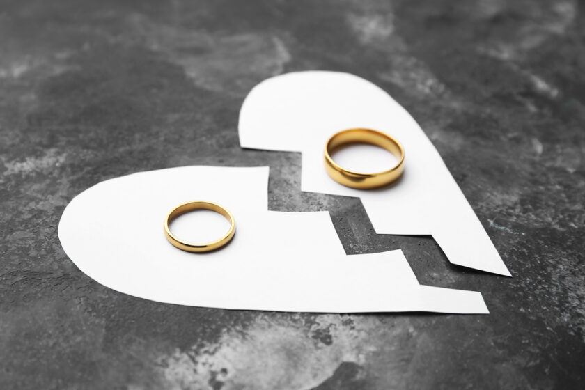 Two gold wedding rings placed on top of a paper heart that is torn in half, symbolising a breakup or divorce, on a dark textured background.