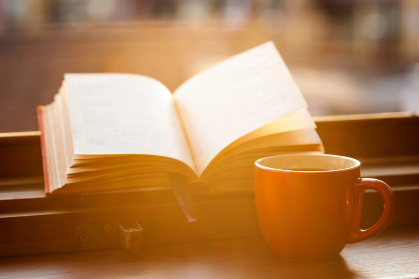 An open book and an orange mug sit on a windowsill, bathed in warm, golden sunlight streaming through the window, creating a cosy and inviting atmosphere.