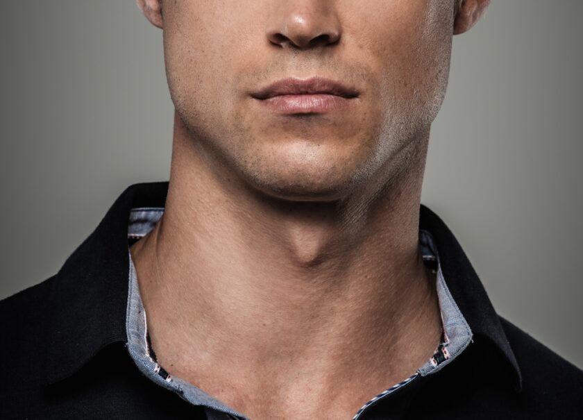 A close-up of the lower half of a man's face and neck, showing his lips, chin, and part of his jawline. He is wearing a dark collared shirt over a light blue inner collar against a neutral background.