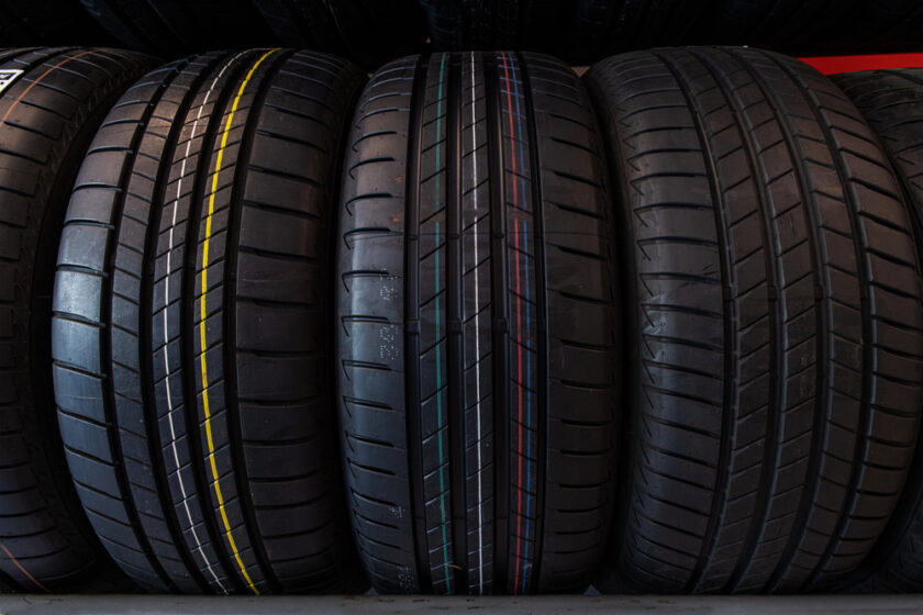 Four black car tyres with deep tread patterns are standing upright side by side. Each tyre has coloured lines (yellow, green, red, blue) running vertically along the treads, against a dark background.