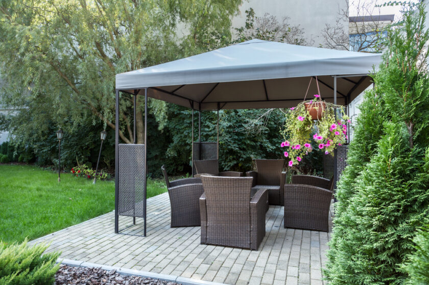 A patio with wicker chairs and a table sits under a grey canopy. Pink flowers hang from the canopy, and the area is surrounded by green trees, bushes, and a neatly mown lawn.