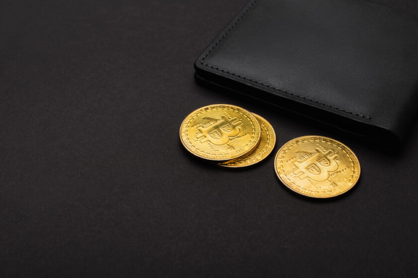 Three gold-coloured Bitcoin coins next to a black leather wallet on a dark surface.