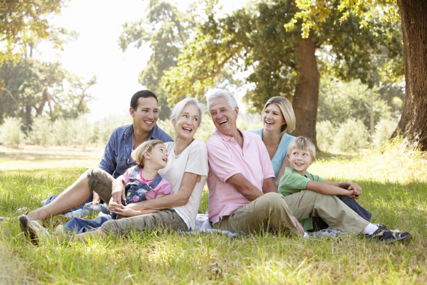 Three adults and three children sit on grass under trees, smiling and laughing together on a sunny day, suggesting a joyful family gathering outdoors.