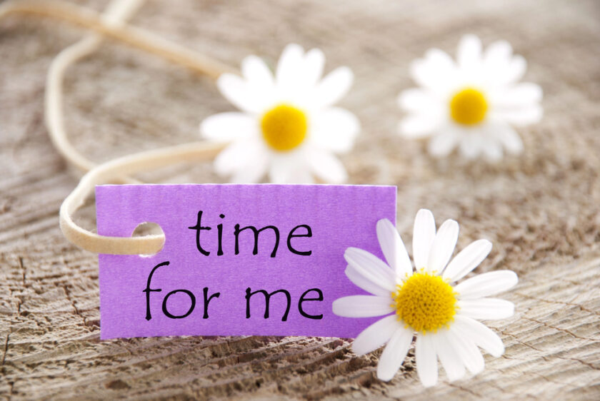 A purple label with the words time for me lies on a wooden surface, surrounded by three white daisies with yellow centres.