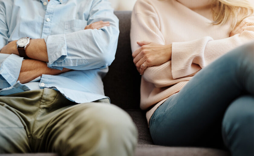 Two people sitting on a sofa with their arms folded, facing slightly away from each other. Their body language suggests frustration or disagreement. Their faces are not visible.