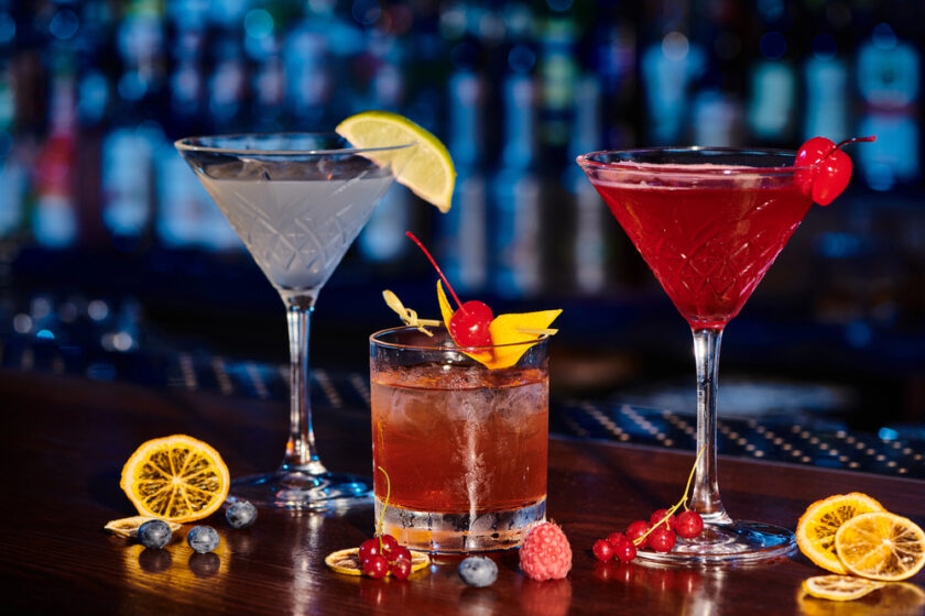 Three colourful cocktails on a bar counter: a clear martini with a lime wedge, a red drink with a cherry, and an amber drink with ice, orange peel, and cherry, surrounded by assorted berries and dried citrus slices.