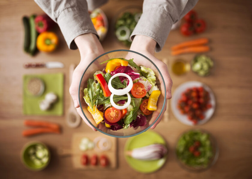 A person holding a bowl of fresh salad with lettuce, cherry tomatoes, yellow bell peppers, and onion slices, above a table filled with assorted vegetables and salad ingredients.