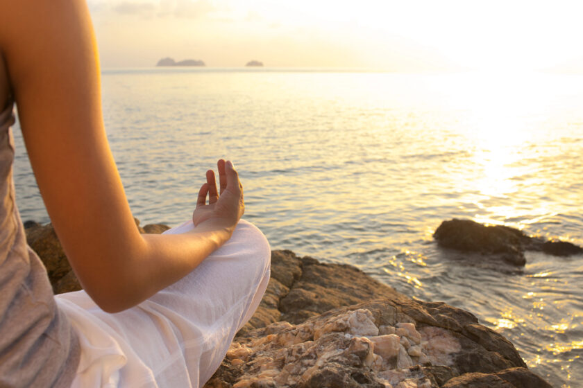 A person sits cross-legged on rocky ground by the sea at sunset, meditating with one hand on their knee and palm facing upwards, wearing light clothing. The calm water and distant islands are visible.