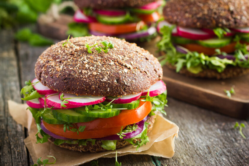 A close-up of a fresh vegetarian sandwich with whole grain bread, sliced radishes, cucumbers, tomatoes, red onions, and leafy greens on parchment paper, with more sandwiches in the background.