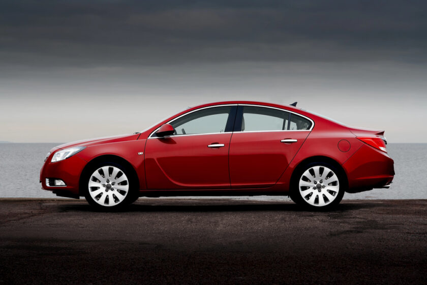 A red saloon car parked on a paved surface near a body of water, with a cloudy sky in the background. The car is viewed from the side, showing its sleek design and white alloy wheels.