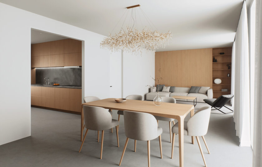 A modern dining and living area features a wooden table with six light grey chairs, a minimalist chandelier, a grey sofa, built-in shelves, and an open kitchen with wooden cabinetry. Natural light fills the space.