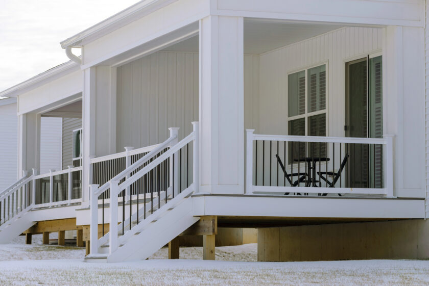 A modern white house with a raised veranda, white railings, and steps. The veranda has a small black table and two chairs. Light snow covers the ground. The scene looks calm and wintry.