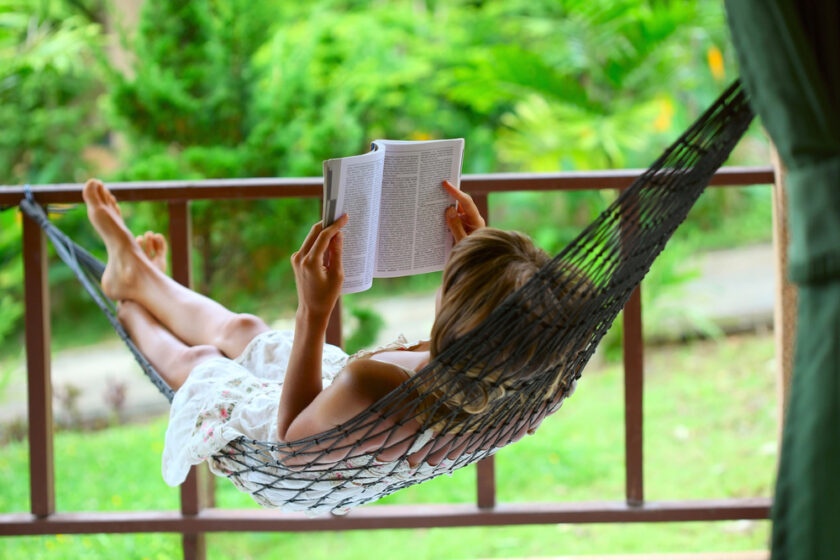A person relaxes in a black hammock on a veranda, reading a book. They are wearing a white dress, and lush green trees and plants fill the background, creating a peaceful outdoor setting.