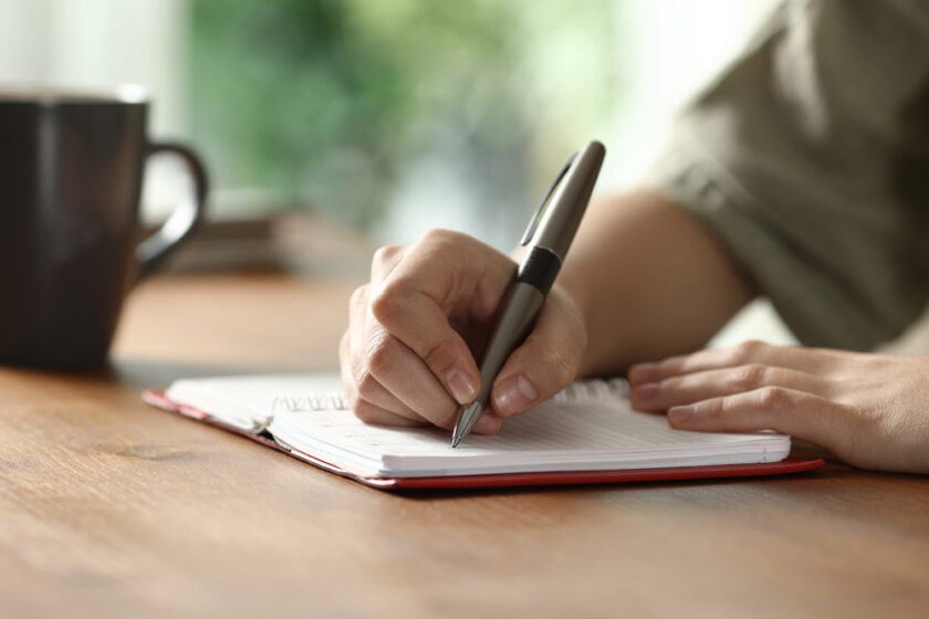 A person writes in a notebook with a pen on a wooden table. A coffee mug sits nearby, and only the person's hands and part of their arm are visible.