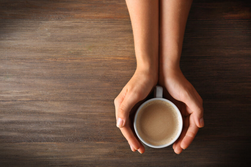 A person’s hands gently cup a white mug filled with coffee, resting on a wooden table, viewed from above.