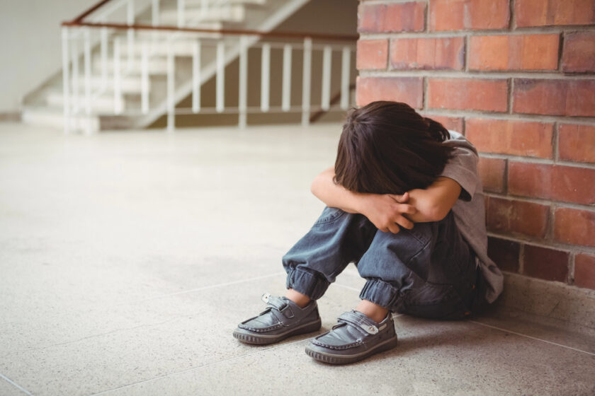 A young child sits on the floor against a brick wall with knees up and head resting on crossed arms, appearing sad or upset in an empty corridor.
