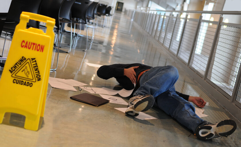 A person lies on the floor near scattered papers in a corridor, appearing to have fallen. A yellow caution sign warning of a wet floor is visible nearby.