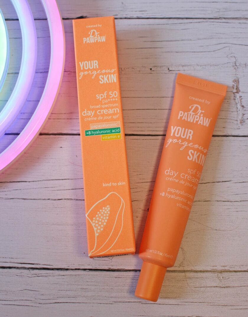 An orange tube and matching box of Dr. PAWPAW Your Gorgeous Skin SPF 50 day cream sit on a light wooden surface beside a glowing neon circle. The packaging highlights papaya, hyaluronic acid, and vitamin C.