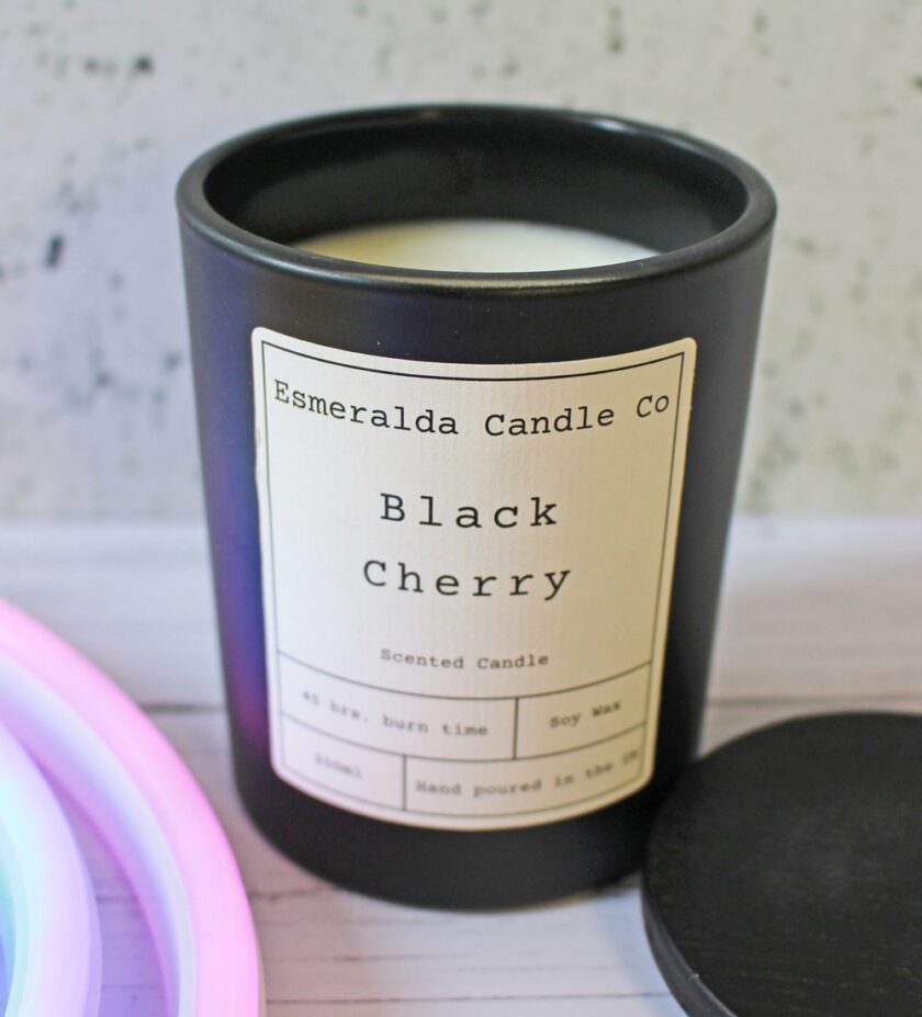 A black candle jar labelled Esmeralda Candle Co Black Cherry sits on a surface with its lid off. The label notes it is a scented soy wax candle, hand poured, with a 45-hour burn time.