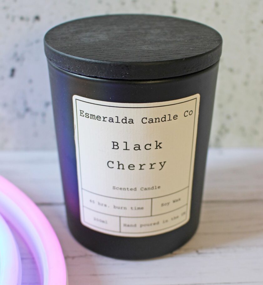 A black candle jar with a wooden lid displays a label reading Esmeralda Candle Co, Black Cherry, Scented Candle, 45 hrs. burn time, Soy Wax, 8oz, Hand poured in the UK. The candle sits on a white wooden surface.
