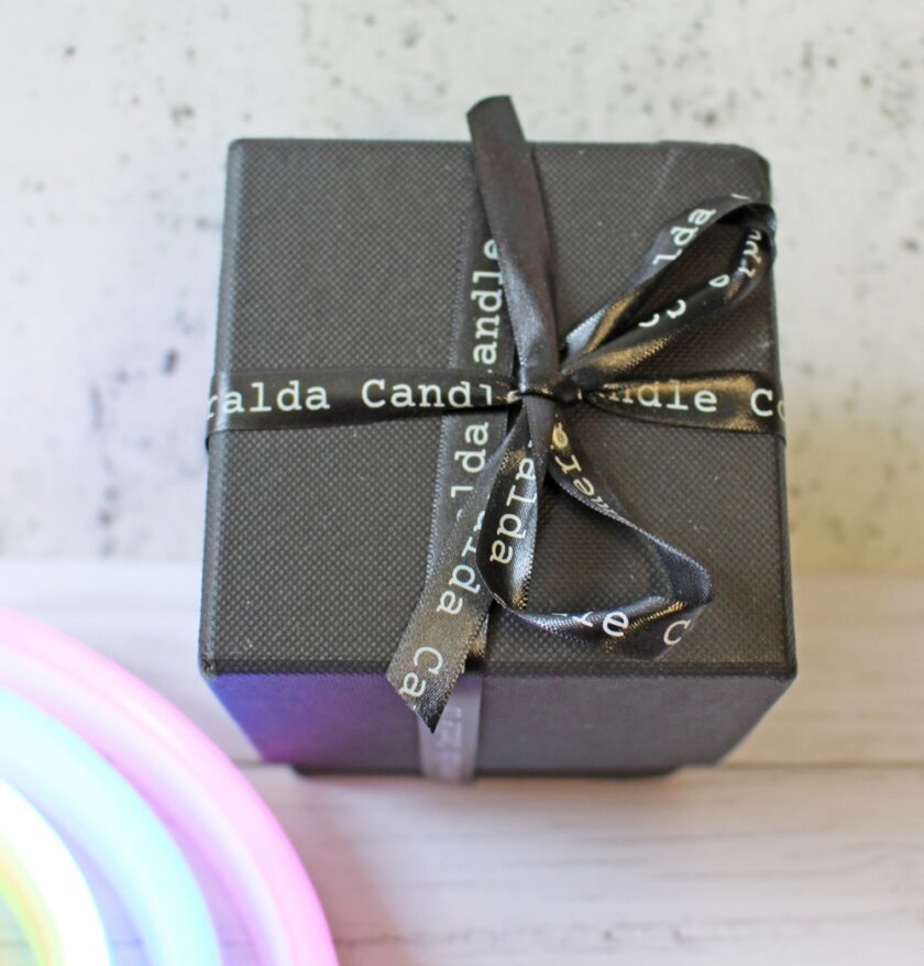 A black gift box tied with a black ribbon that has white text reading Candle, placed on a light wooden surface with a blurred, colourful light in the corner.