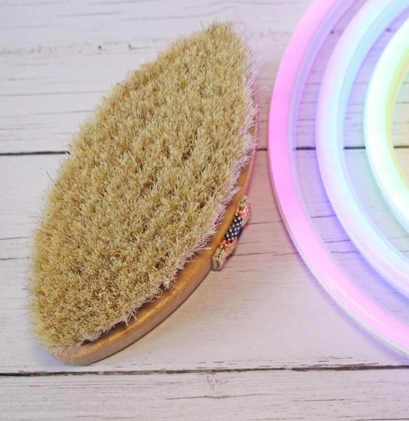 A dry body brush with bristles and a beaded handle rests on a white wooden surface next to pastel-coloured neon lights.