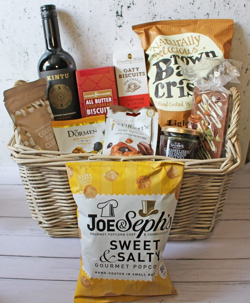 A wicker hamper filled with assorted gourmet snacks, including wine, popcorn, crisps, biscuits, nuts, chocolate, and a jar of preserves, displayed on a light wooden surface with a white background.