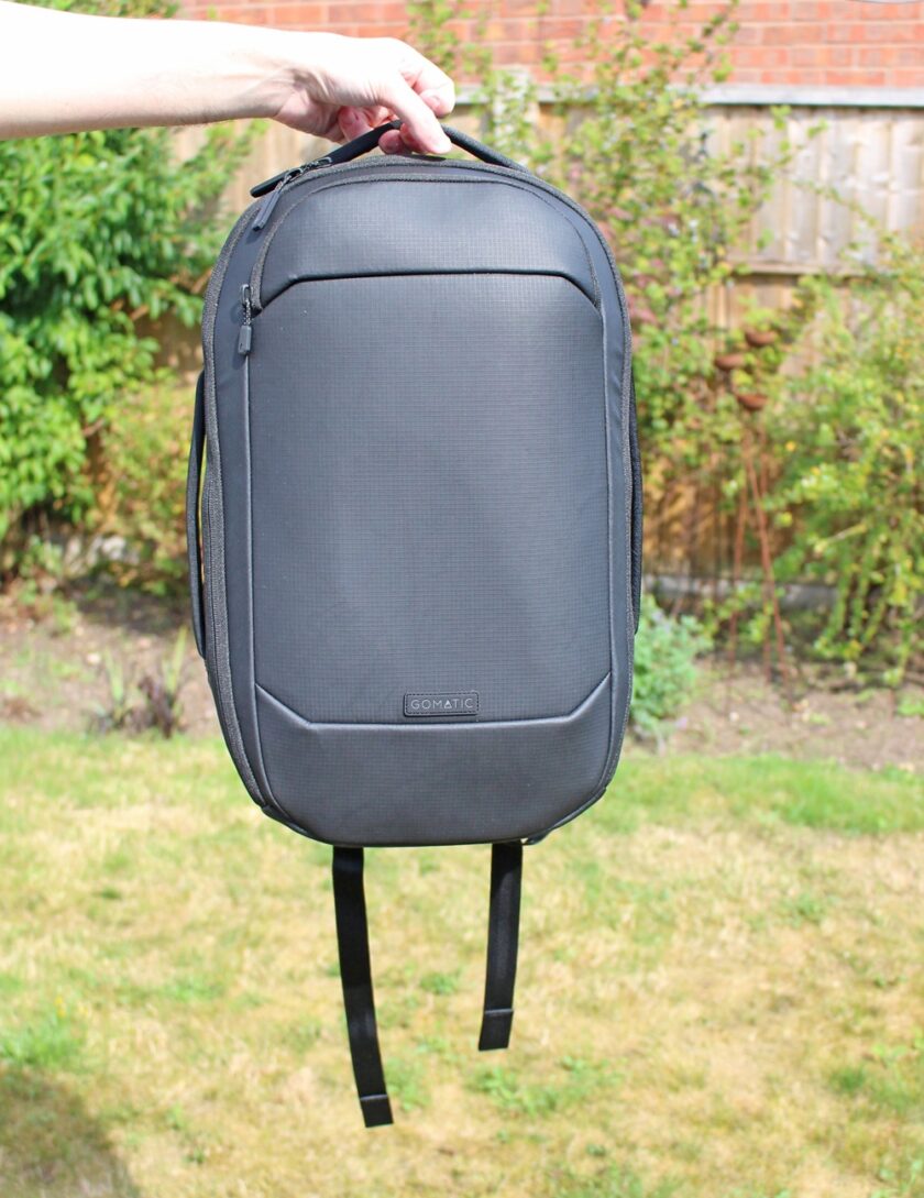 A hand holds up a sleek, black rucksack with a rectangular shape and minimal design, outdoors in a grassy garden with bushes and a wooden fence in the background.
