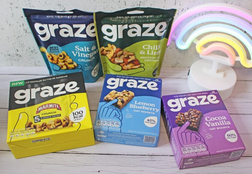 Five Graze snack packs—Salt & Vinegar, Chilli & Lime, Marmite, Lemon Blueberry Oat Boosts, and Cocoa Vanilla Oat Boosts—are displayed on a white wooden surface next to a colourful neon rainbow light.