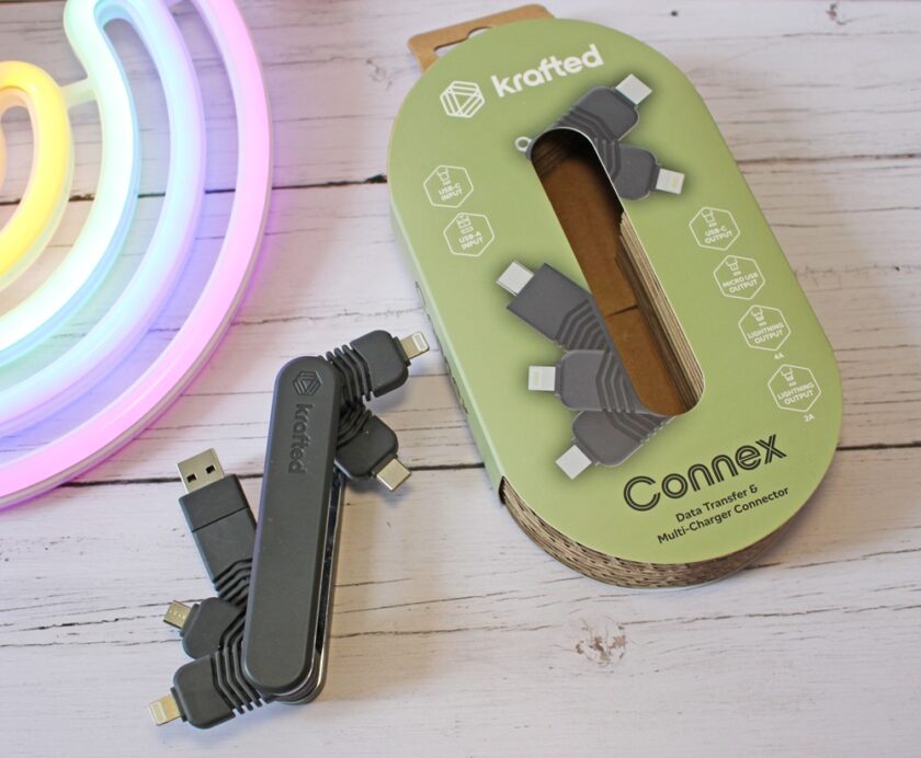 A multi-charging connector with USB and various charging tips lies next to its green Krafted Connex packaging on a white wooden surface, with part of a colourful neon light visible in the top left corner.