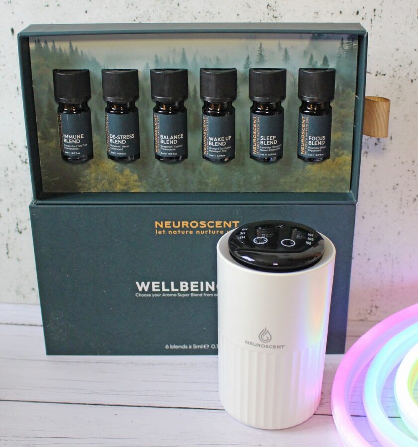A box containing six small essential oil bottles labelled with different blends is displayed above a white electric diffuser. The box and diffuser are branded Neuroscent. Part of a colourful light is visible on the right.