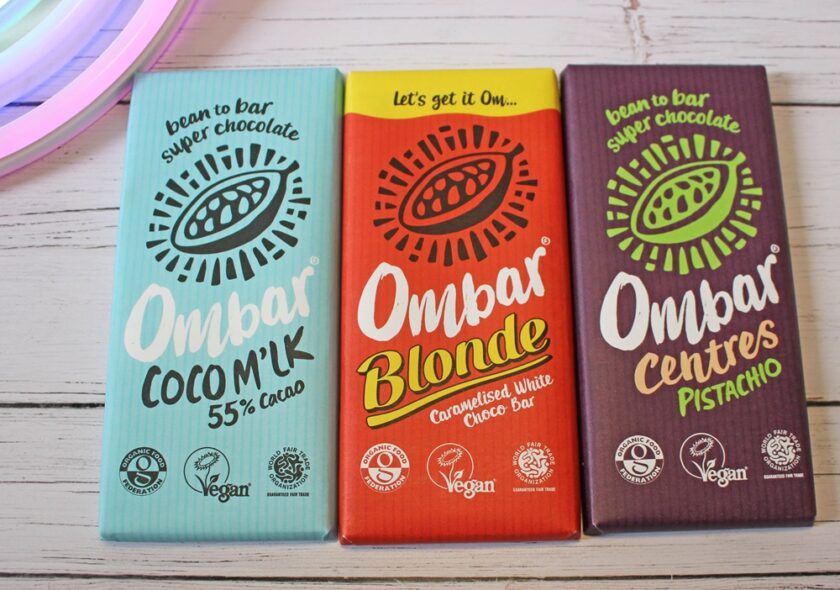 Three Ombar chocolate bars are displayed on a white wooden surface: Coco Mylk (blue wrapper), Blonde Caramelised White (orange wrapper), and Centres Pistachio (purple wrapper). All are labelled vegan.