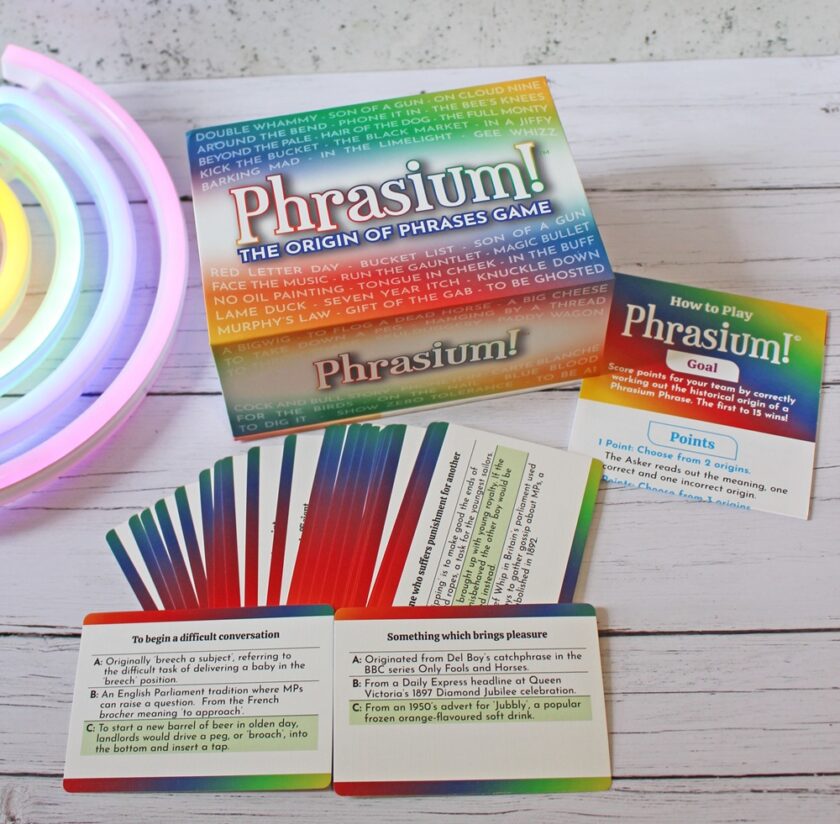 A board game called Phrasium! The Origin of Phrases Game is displayed on a white wooden surface with neon lights in the corner. The game includes colourful cards, instructions, and a box.