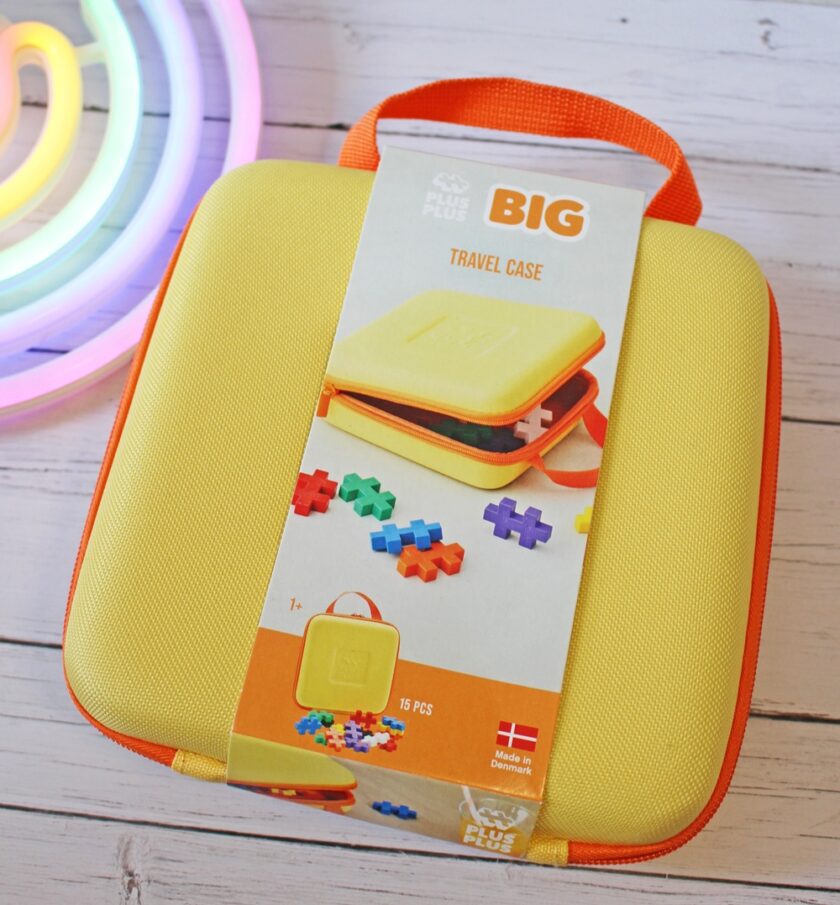 A yellow and orange Plus-Plus BIG travel case with a handle, shown in packaging with colourful interlocking pieces, placed on a white wooden surface next to a rainbow-shaped light.