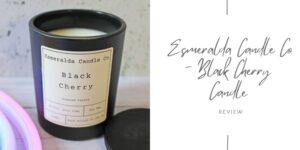 A black Esmeralda Candle Co. candle labelled Black Cherry sits on a light surface with its lid beside it. Next to the candle is text that reads Esmeralda Candle Co - Black Cherry Candle Review.