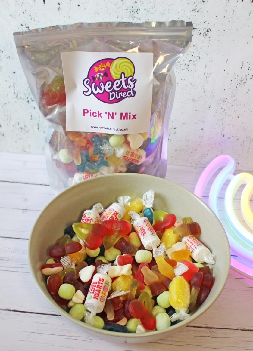 A bowl of assorted colourful sweets and confectionery sits in front of a resealable bag labelled Sweets Direct Pick N Mix. The background includes a neon light in the shape of a rainbow.