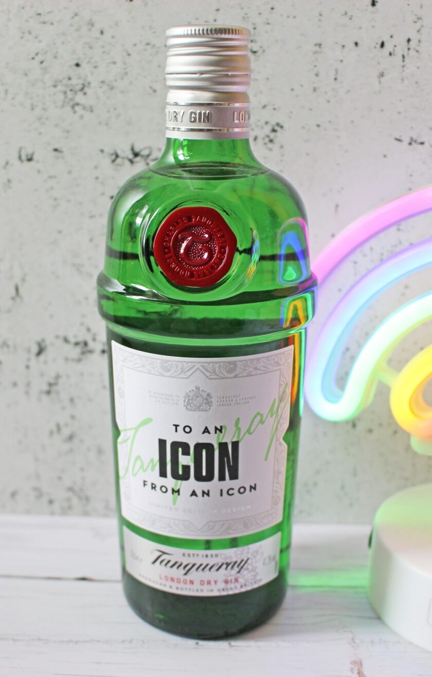 A green bottle of Tanqueray London Dry Gin with a special label that reads To an Icon from an Icon, placed on a white surface next to a colourful, neon rainbow light.