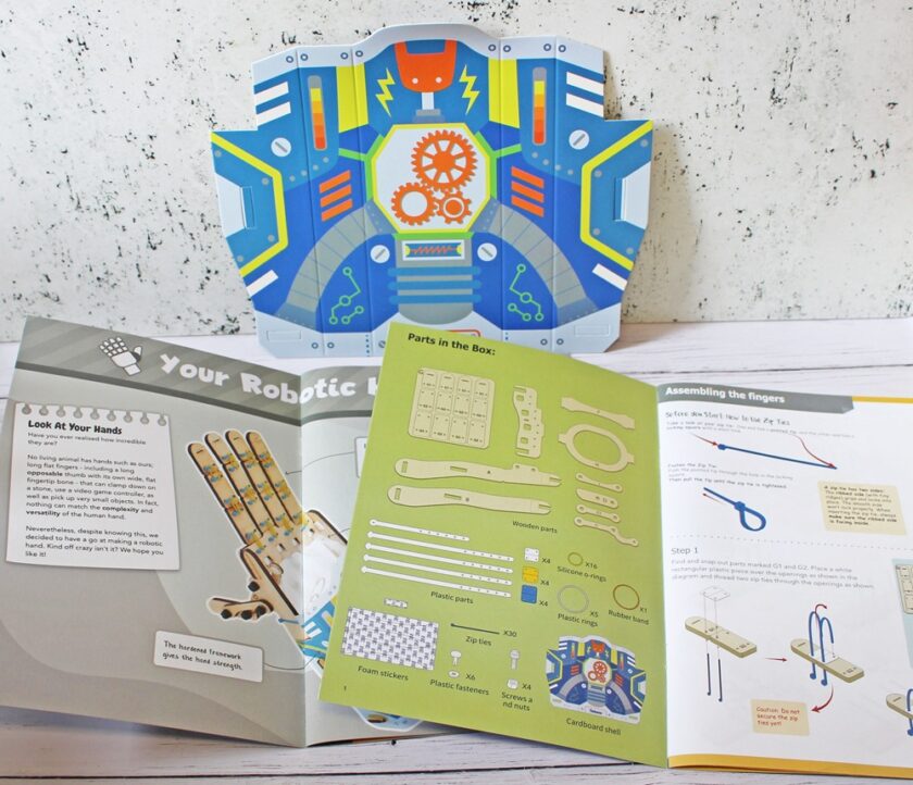 An open STEM activity kit displays illustrated instruction booklets, cut-out cardboard pieces, and diagrams for assembling a robotic hand, with colorful gears and parts shown on the pages.
