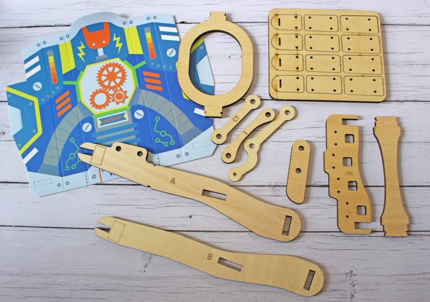 A DIY robitic hand kit is laid out on a white wooden surface, featuring colorful printed cardboard pieces and laser-cut wooden parts with various shapes and slots, ready to be assembled.