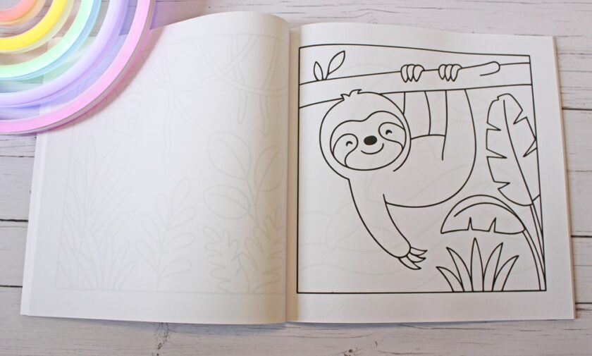 An open colouring book shows a page with an outlined drawing of a smiling sloth hanging from a tree branch. The book rests on a white wooden surface beside a colourful, curved object.
