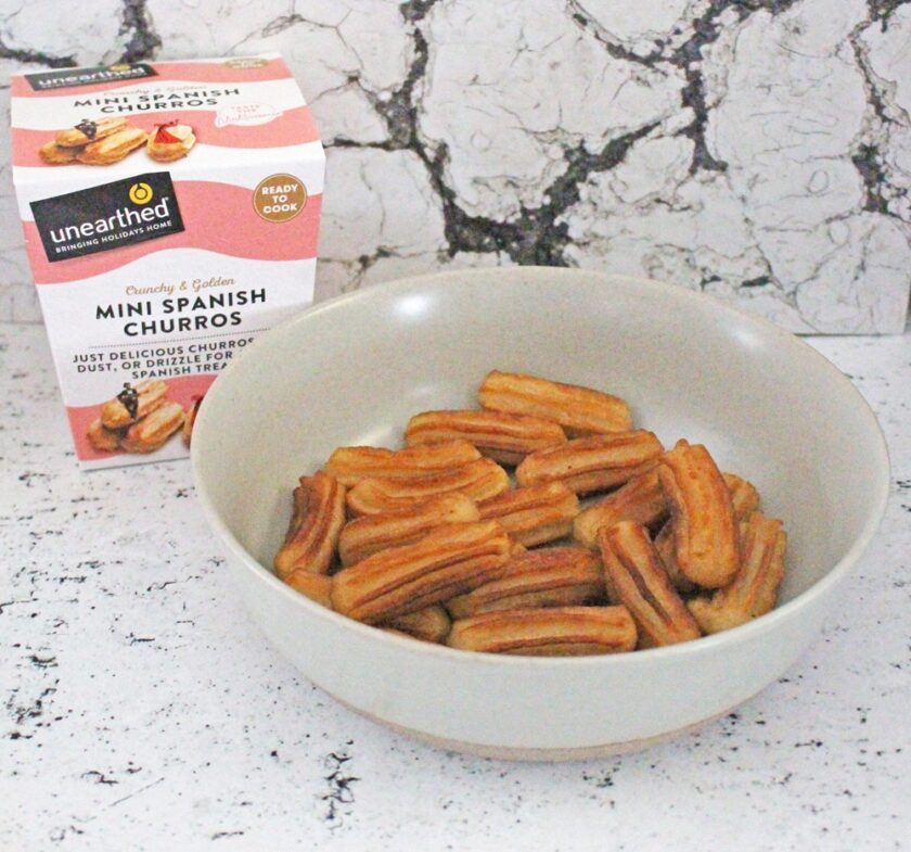 A bowl filled with mini Spanish churros sits on a white, marbled surface. Behind the bowl is a box labelled Mini Spanish Churros with images of churros on the packaging.