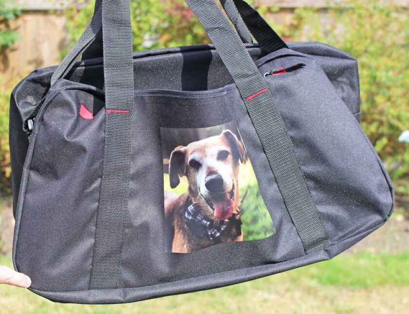 A black duffle bag with a photo of a brown dog wearing a checked bandana printed on the front pocket, shown outdoors with greenery in the background.