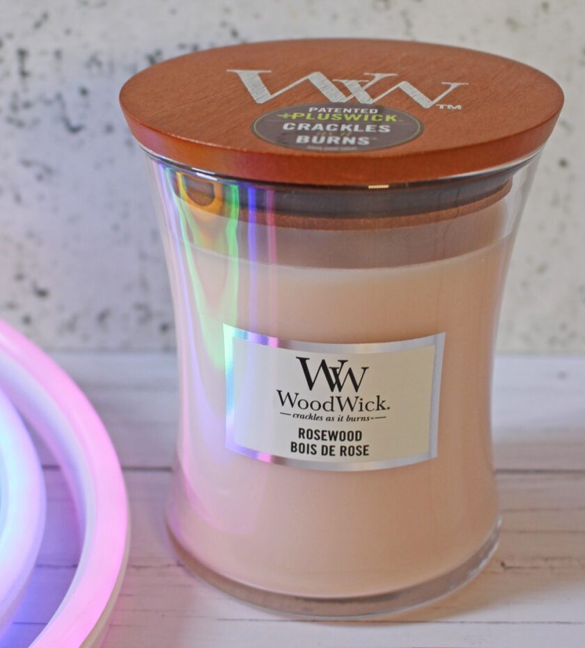 A WoodWick Rosewood scented candle with a wooden lid sits on a light surface. The candle is in a clear glass jar with a white label, and soft rainbow light reflects on the glass.