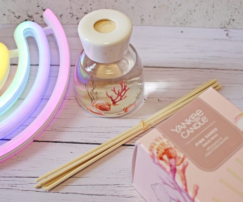 A bottle of Yankee Candle Pink Sands reed diffuser with decorative sea shell and coral designs, wooden reeds, and a pastel neon light on a white wooden surface.