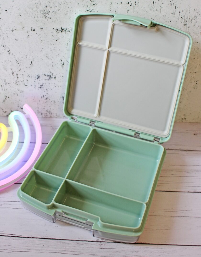 A pastel green lunchbox with four compartments sits open on a light wooden surface. A multicoloured neon rainbow light decorates the background near a white, textured wall.